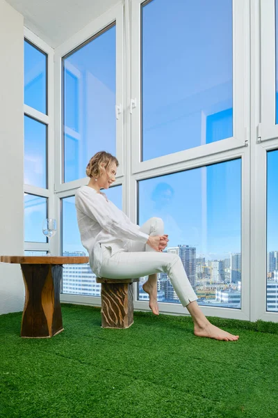 Barefoot woman sits on the balcony of her new apartment and admires the view of the city from the window