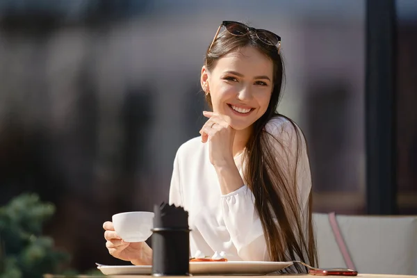 Cute young woman with cup of cappuccino in hand looks straight and smiles