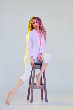 Menekşe ve sarı ışıklarla aydınlatılmış beyaz elbiseli genç bir kadının tam boy portresi.