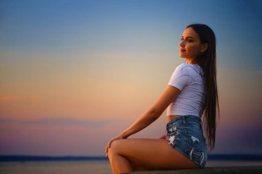 Young woman sitting in profile on a pier and watching the sunset
