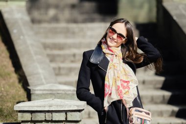 Paltolu ve deri pantolonlu mutlu genç kadın rüzgarlı bir günde saçlarını düzeltiyor.