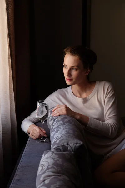 Mujer bonita sentada cerca de la ventana en la oscuridad con un vaso de vino —  Fotos de Stock