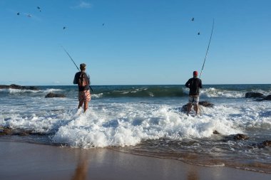 Salvador, Bahia, Brezilya - 25 Nisan 2021: Salvador, Bahia 'daki Rio Vermelho plajında kayaların üzerinde silüet halinde balıkçılar. İyi balıkçılık günleri..