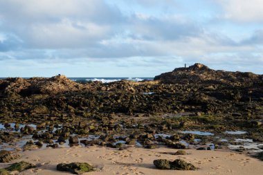 Mavi gökyüzü ve deniz kayaların üzerinde ve kumsalda kum. Dalgalar kayalara çarpıyor ve kumsalda kum var..