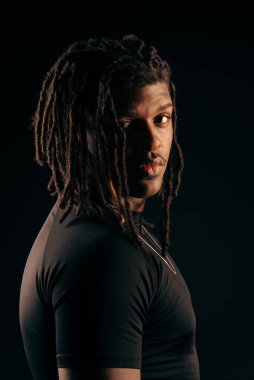 Siyah tişörtlü, rastalı yakışıklı bir adam stüdyoda kameraya bakıyor. Siyah bir arka planda izole edilmiş. Fotoğraf modeli.