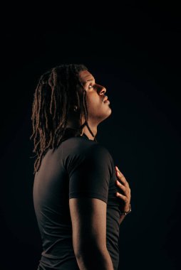 Siyah tişörtlü, rastalı yakışıklı bir adam stüdyoda kameraya bakıyor. Siyah bir arka planda izole edilmiş. Fotoğraf modeli.