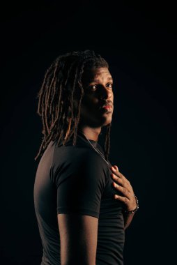 Siyah tişörtlü, rastalı yakışıklı bir adam stüdyoda kameraya bakıyor. Siyah bir arka planda izole edilmiş. Fotoğraf modeli.