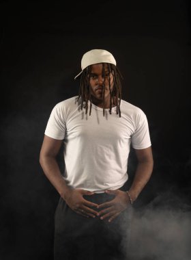 Rastalı ve beyaz şapkalı yakışıklı bir genç stüdyoda fotoğraf çekimi sırasında kameraya poz veriyor. Dumanlı bir arkaplanda izole edilmiş.