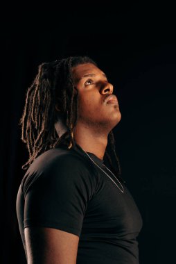 Siyah tişörtlü, rastalı yakışıklı bir adam stüdyoda kameraya bakıyor. Siyah bir arka planda izole edilmiş. Fotoğraf modeli.