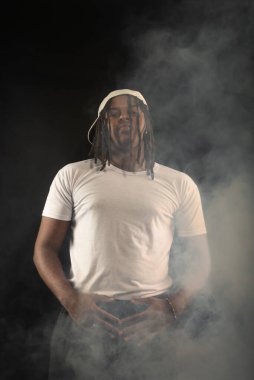 Rastalı ve beyaz şapkalı yakışıklı bir genç stüdyoda fotoğraf çekimi sırasında kameraya poz veriyor. Dumanlı bir arkaplanda izole edilmiş.