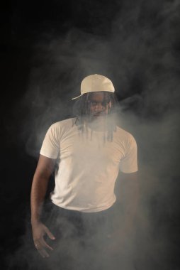Rastalı ve beyaz şapkalı yakışıklı bir genç stüdyoda fotoğraf çekimi sırasında kameraya poz veriyor. Dumanlı bir arkaplanda izole edilmiş.