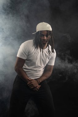 Rastalı, şapkalı ve beyaz gömlekli bir adam stüdyoda dumanlı bir arka planda kameraya poz veriyor. Kendinden emin ve ciddi bir genç adam..