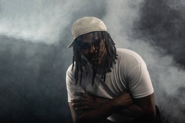 Rastalı, şapkalı ve beyaz gömlekli bir adam stüdyoda dumanlı bir arka planda kameraya poz veriyor. Kendinden emin ve ciddi bir genç adam..