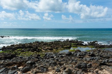Kaya oluşumları ve deniz kenarındaki doğal havuzlarla panoramik bir sahil manzarası. Mavi okyanus, parçalı bulutlu bir gökyüzünün altında ufka doğru uzanıyor ve tropikal kıyıların tipik sakin ve doğal atmosferini oluşturuyor..