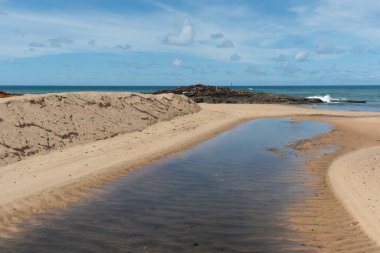 Küçük bir dere ya da tatlı su birikintisini gösteren bir sahil manzarası ve altın kumun içinden akan lağım okyanusa giden bir yol yaratıyor..