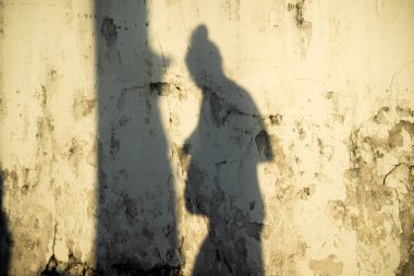 Ayakta duran, muhtemelen telefon kullanan ya da elinde bir şey tutan eski, yıpranmış, açık renkli bir duvara yansıyan uzun bir silüet..