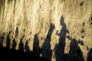 Bir grup insan ve motosiklet eski, kaba, bej sarısı bir duvara yansıtılıyor. Yoğun, düşük güneş ışığı güçlü bir kontrast ve drama yaratır..