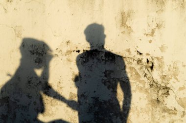 İki farklı gölge dokulu, sarımsı bir duvara yansıyor. Motosikletlilerden biri kask takıyor, diğeri yaya yürüyüşü yapıyor..