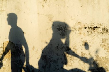 İki farklı gölge dokulu, sarımsı bir duvara yansıyor. Motosikletlilerden biri kask takıyor, diğeri yaya yürüyüşü yapıyor..