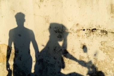 İki farklı gölge dokulu, sarımsı bir duvara yansıyor. Motosikletlilerden biri kask takıyor, diğeri yaya yürüyüşü yapıyor..