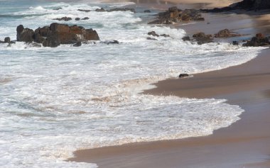 Dalganın kırılma ve geniş bir beyaz köpük tabakasına dönüşmesinin ayrıntıları ıslak kum şeridinde ilerlerken, arka planda kayalar açıkta kalır..