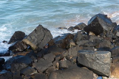 Kumsalın kenarındaki karanlık, siyah, ıslak taşlar ve kayalar güneşli bir günde köpüklü, beyaz deniz dalgaları tarafından vuruluyor..