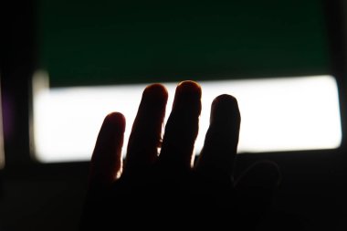 Ön planda açık, uzanmış bir el, siluet içinde, bir pencereden veya açılıştan gelen güçlü bir ışığın arka planına karşı, yeşil ve beyaz alanları ile.