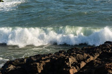 Beyaz köpüklü bir dalganın kayalık kıyıya vuruşunun yan görüntüsü.