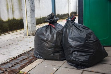 İki büyük, siyah çöp torbası yeşil bir çöp tenekesinin ve paslı bir ızgaranın yanında bağlı duruyor..