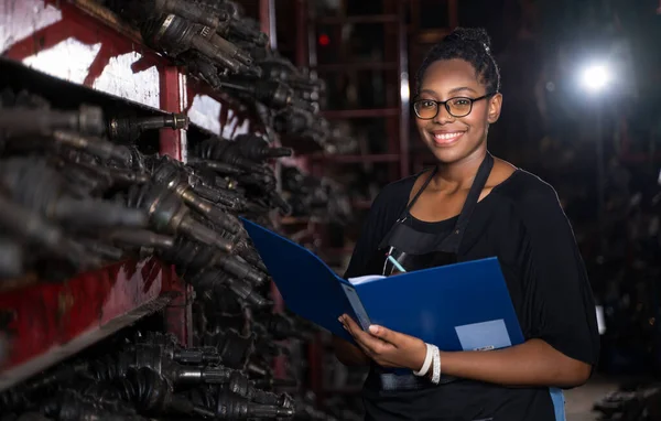 A black woman, business owner or employee in a factory engine parts. Holding the file to check the product list. Smiling and looking at the camera n Business onwner and worker concept