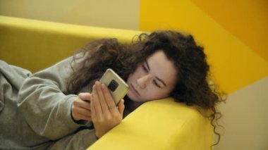 Kız sarı bir kanepede uzanıyor ve cep telefonundan son haberleri okuyor. Arka plandaki duvarda sarı ve turuncu renklerin geometrik deseni