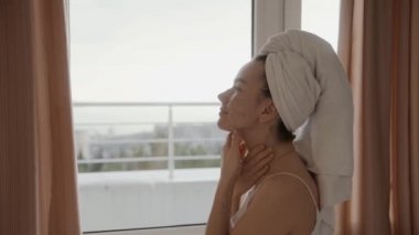 Beyaz havluyla cildinde sorun olan genç bir kız yüz derisine krem sürüyor. Sabahları yüz masajı tazeleniyor. Yanaklarında deri döküntüleri ve sivilceler var. Yüz bakımı ürünü uygulanıyor