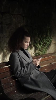 Parktaki bankta oturan ve telefona bakan bin yıllık bir kızın dikey yavaş görüntüsü. Bir iletiyi yazdırır ya da bir uygulama kullanır. Şehir kıyafetleri içinde, gri paltolu ve kıvırcık saçlı genç bir kadın. 