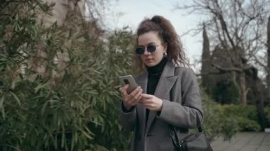 A young woman in a gray coat and sunglasses walks along a city street near a hedge. Curly hair. Conversation on a mobile phone. Everyday calling a friend or chatting with relatives 