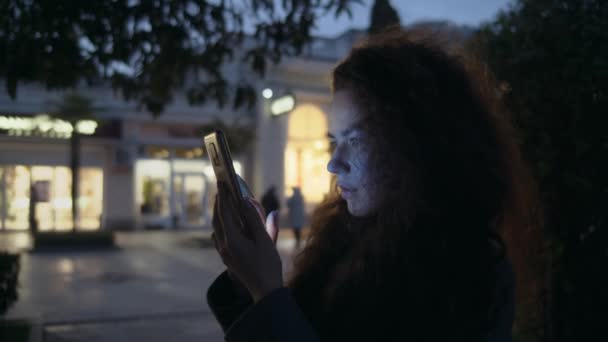 傍晚时分 一位身穿外套的卷发女孩在街上检查她的手机 商店橱窗在后面点着灯 在网上购物或在城市里到处走走 — 图库视频影像