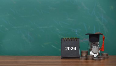 Çalışma hedefleri, 2026 masa takvimi, mezuniyet şapkası ve bozuk para yığınıyla dolu cam şişe. Eğitim, öğrenci kredisi, burs, 2026 yılının okul harcı için para biriktirme kavramı