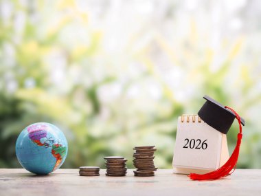 Hedeflere çalış. Mezuniyet şapkası, bozuk para yığını ve Dünya küresi olan 2026 masa takvimi. 2026 yılında eğitim, öğrenci kredisi, burs, öğrenim ücreti için para biriktirme kavramı