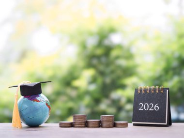 Hedeflere çalış. Mezuniyet şapkalı dünya küresi, 2026 masa takvimi ve bir yığın bozuk para. 2026 yılında eğitim, öğrenci kredisi, burs, öğrenim ücreti için para biriktirme kavramı