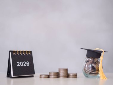 Çalışma hedefleri, 2026 masa takvimi, mezuniyet şapkası ve bozuk para yığınıyla dolu cam şişe. 2026 yılında eğitim, öğrenci kredisi, burs, öğrenim ücreti için para biriktirme kavramı