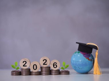 Mezuniyet şapkalı dünya küresi, 2026 numaralı tahta blok ve bozuk para yığınında büyüyen bitki. 2026 yılında eğitim, öğrenci kredisi, burs, öğrenim ücreti için para biriktirme kavramı.