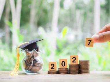 Mezuniyet şapkalı para yığınının üzerinde 2027 numaralı tahta bloklar var. 2027 yılında eğitim, öğrenci kredisi, burs, öğrenim ücreti için tasarruf kavramı