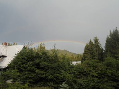Ilıman bir dağ köyünün üzerinde parlak bir gökkuşağı beliriyor. Yoğun ladin ormanlarıyla çevrili. Metal çatılar ve her yer yemyeşil olmuş bir baca. Karanlık fırtına bulutları ormanlık bir tepenin üzerinden yükselirken..