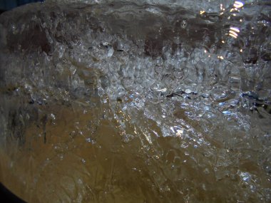 Hava kabarcıkları ve ince çatlaklarla dolu bir buz kütlesine yakın çekim. Kristal yüzey, düşük ışıkta parlak parlak ışıklar ve sıcak yansımalar göstererek soyut bir model oluşturur..