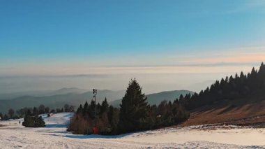 Mevsim dışında suni kar ve kozalaklı orman ile kayak yamacında dağ manzarası ve ufukta güneş var..
