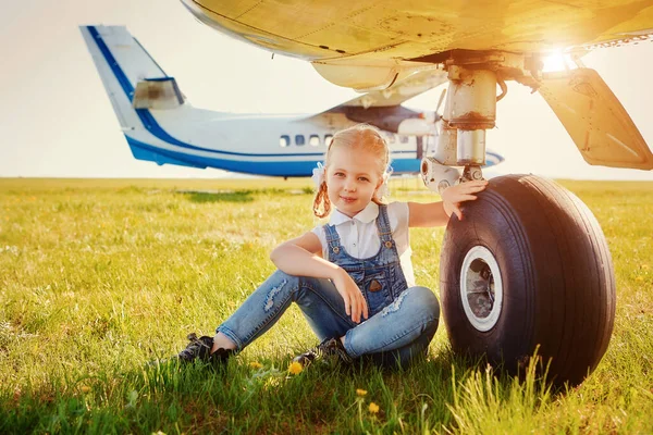 Linda niña sentada cerca de las ruedas del tren de aterrizaje de un avión y soñando con viajar y volar en un avión — Foto de Stock