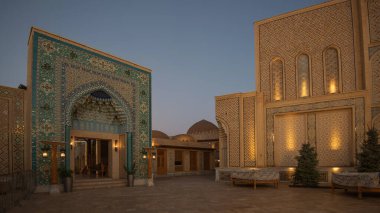 Semerkant, Özbekistan 'ın İpek Yolu kompleksindeki avluda, akşamüstü erken saatlerde turkuaz mozaik kemerler, geleneksel kubbeler, kemerli pencereler, banklar ve aydınlatılmış duvarlar yer alıyor..
