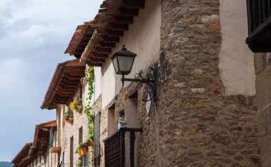 Rustic stone buildings line a narrow street in a charming Spanish village. The weathered facades feature hanging flower pots and wrought iron balconies.