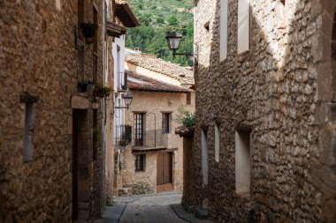 A narrow cobblestone street winds through a charming Spanish village, flanked by rustic stone buildings. Old-fashioned lanterns adorn the facades, creating picturesque atmosphere.