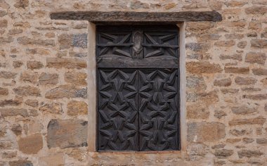 An ancient wooden window set in a weathered stone wall, located in a historic village in Spain. The detailed geometric patterns of the woodwork contrast beautifully with the rough texture of the stone