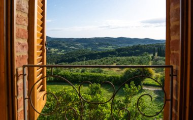 View through a rustic wooden window reveals rolling Tuscan hills with olive groves, cypress trees, and lush vegetation. Wrought iron balcony railing frames the picturesque countryside scene.